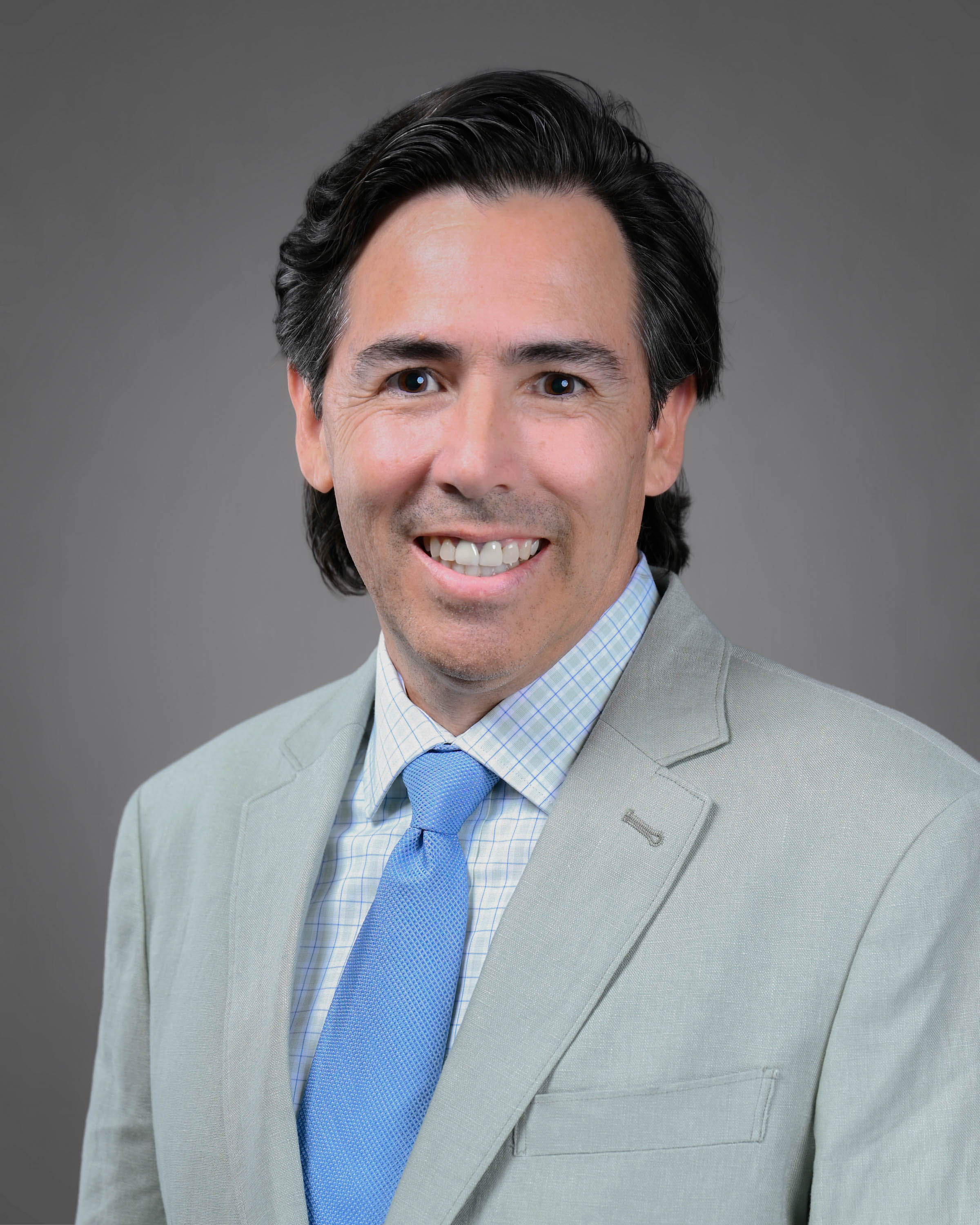 Smiling man with longer dark hair in a grey suit with a checkered shirt and blue tie.